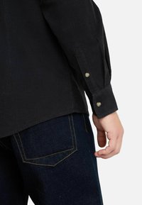 Camicia di velluto a coste nera con polsini abbottonati, abbinata a jeans in denim scuro con cuciture gialle sulle tasche.