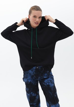 Černá mikina s kapucí a zelenými tkanicemi, volný střih; spárována s modrými cargo kalhotami s tie-dye vzorem a několika kapsami.