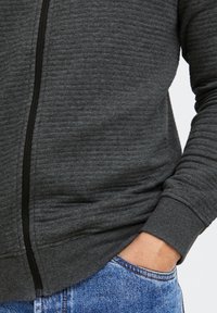 Sudadera con cremallera de color gris oscuro, con un aspecto texturizado y cremallera negra. Presenta puños acanalados y un bolsillo frontal, combinada con jeans de mezclilla azul.
