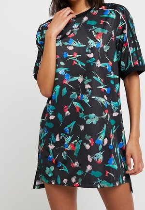 Vrouw draagt een zwart Adidas-jurkje met kleurrijke bloemenprints, korte mouwen en drie witte strepen op de schouders.