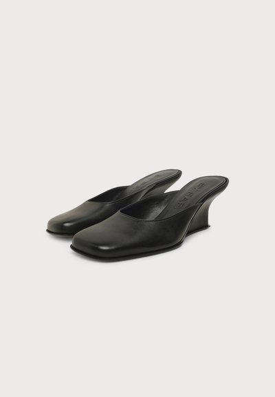 Paire de chaussures mules en cuir noir à enfiler avec des talons compensés bas et incurvés sur fond blanc.