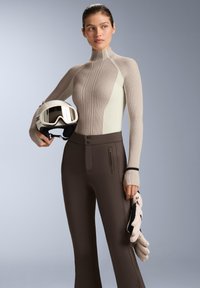 Tenue de ski comprenant un haut à manches longues côtelé beige et vert clair, un pantalon de ski marron et un casque blanc tenu à la main. Des gants texturés sont inclus.