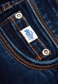 Tumman sininen farkkukangas, jossa on oranssit tikkaus, metallinen neppari ja valkoinen etiketti, jossa lukee "Superdry" sinisellä tekstillä.