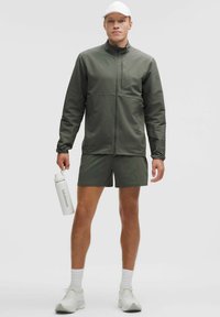 Grøn atletisk jakke med et tekstureret mønster, parret med matchende shorts. Bærer en hvid cap og holder en hvid vandflaske. Hvide sneakers.