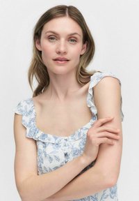 Kwiatowa sukienka w białym kolorze z niebieskimi wzorami, z falbaniastymi ramiączkami i kwadratowym dekoltem, z detalami w postaci guzików na przodzie. Miękka tekstura materiału.