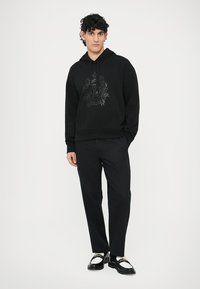 Black hoodie with a tonal graphic design, worn with black trousers and black and white footwear. Smooth fabric texture and relaxed fit.
