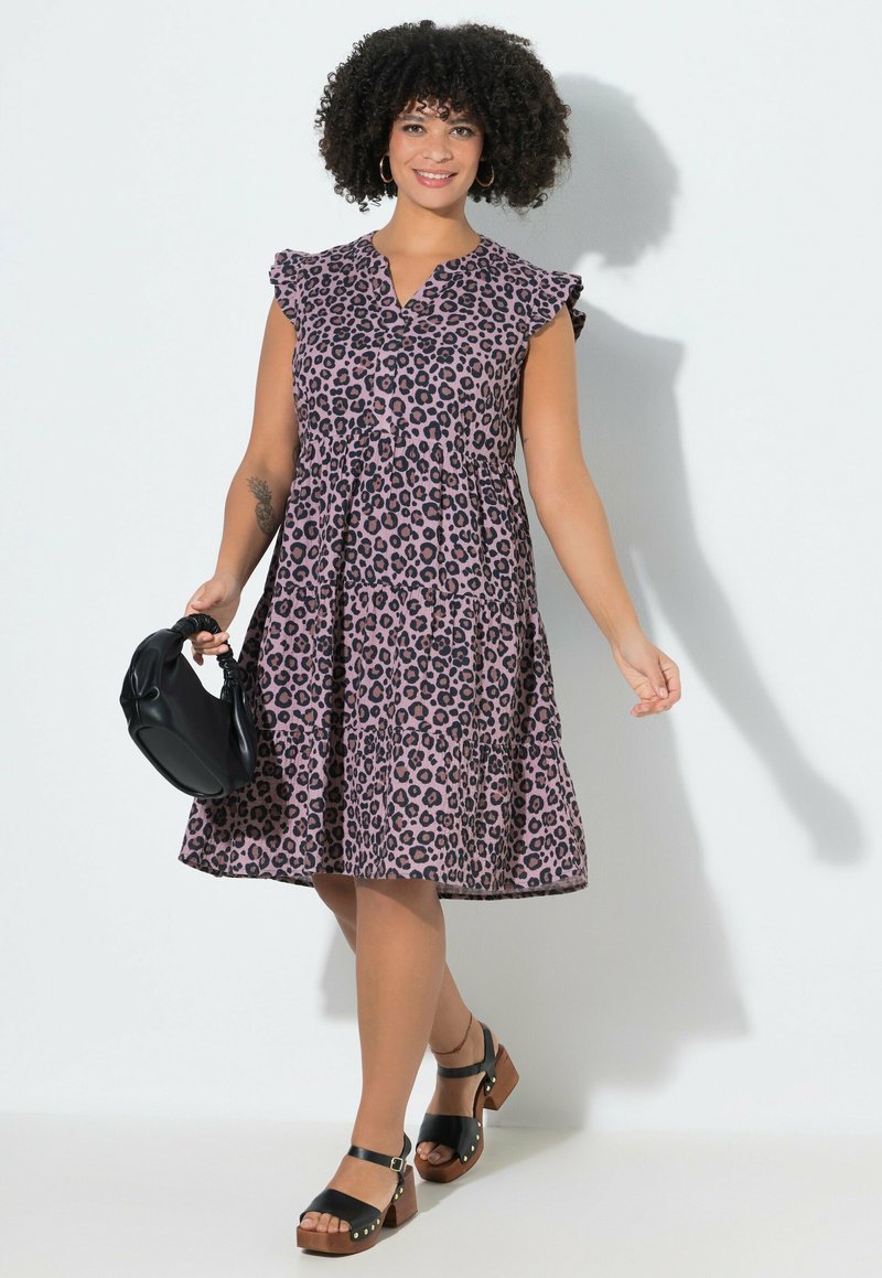 Donna con capelli ricci che indossa un vestito rosa senza maniche con stampa leopardata, sandali neri e tiene una piccola borsa nera.