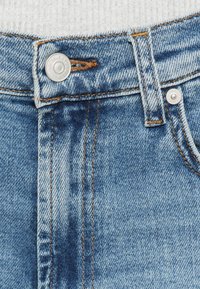 Helelein sinised teksapüksid, millel on nööpkinnitus, kontrastne õmblus ja tekstuuriga kangas, lähivõte vöökohast.