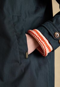 Ręka osoby w ciemnoniebieskiej kieszeni płaszcza, pokazująca dzianinowy mankiet w pomarańczowo-białe paski oraz brązowe guziki na rękawie.