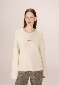Maglione beige con orlo a coste, caratterizzato dalla parola "oui" ricamata in rosso. Indossato sopra pantaloni grigi con stampa leopardata. Tessuto morbido, vestibilità ampia.