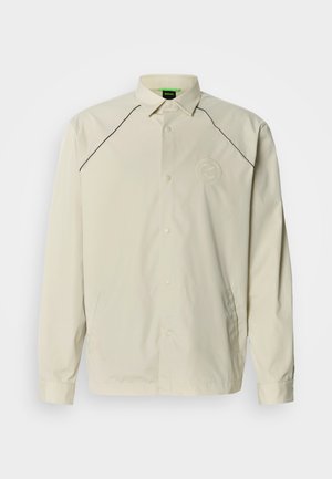 Bež lahka jakna z ostro ovratnico, dolgimi rokavi, črnim zaključkom ob ramenih in okrogo logotipom na levi strani prsnega koša.