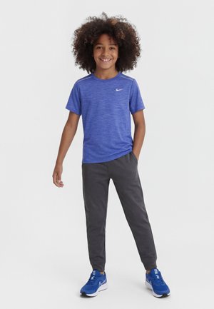 Lächendes Kind mit lockigem Haar, das ein blaues Nike-T-Shirt, dunkle Jogginghosen und blaue Sneakers trägt und mit der Hand in der Tasche steht.