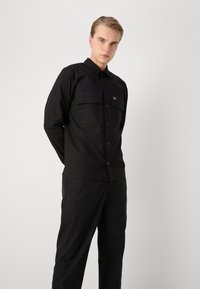 Μαύρο πουκάμισο με κουμπιά, με δύο τσέπες στο στήθος, μακριά μανίκια και κα pointed κολάρο. Κατασκευασμένο από μαλακό, ματ ύφασμα με κεντημένο λογότυπο.