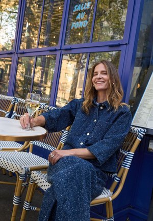 Femme en tenue bleue assise à une table de café en extérieur, tenant un verre de vin blanc, souriante avec une fenêtre bleue et un menu derrière elle.