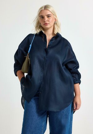 Donna bionda che indossa una camicia oversize blu navy con bottoni e dei jeans blu, porta una piccola borsa oliva con tracolla blu sulla spalla.