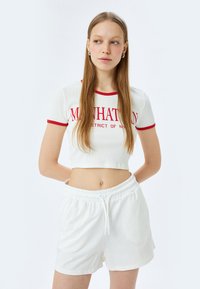 Witte geribde crop top met rode bies, met de tekst "MANHATTAN DISTRICT OF NY"; gecombineerd met witte shorts met elastische taille.