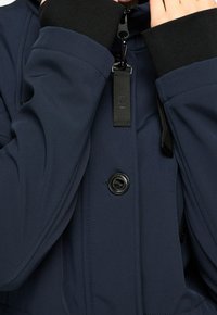 Marinblå isolerad jacka med hög krage, svarta ribbade manschetter, knäppning fram med knappar och justerbara remmar med metalldetaljer.