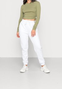 Olijfgroen langemouwen top met gathered zijkanten, gecombineerd met losse witte joggers en witte sneakers, op een lichtgrijze achtergrond.
