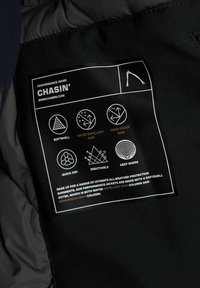 Etiqueta negra de cuidado con iconos de repelencia al agua, a prueba de viento, de secado rápido, transpirabilidad y calidez. El texto incluye la marca "CHASIN'."