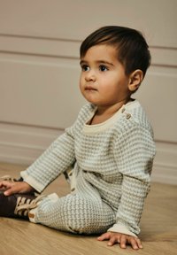 Un tout-petit avec des cheveux foncés portant une tenue en tricot beige et bleue à motifs, assis sur un sol en bois, regardant sur le côté.