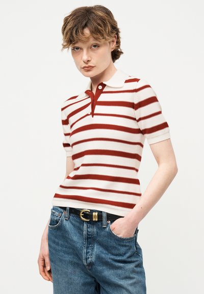 Jeune personne portant un polo à manches courtes blanc à rayures rouges et un jean bleu avec une ceinture noire, debout avec une main dans la poche.