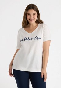 Camiseta blanca de manga corta con cuello en V y "La Dolce Vita" impresa en azul marino. Textura suave, corte holgado y mangas con dobladillo. Combinada con pantalones azul marino.