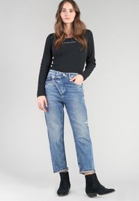 Haut noir à manches longues avec un effet de côtes texturé, associé à un jean en denim déchiré bleu clair et des bottines noires, présenté sur un fond gris.