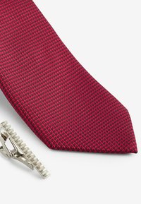 Red tie with a textured pattern, accompanied by a silver tie clip. The tie features a narrow design and a pointed tip.
