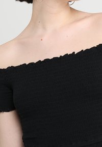 Černý top bez ramínek vyrobený z texturované látky s řasenými okraji a elastickým výstřihem pro přiléhavý vzhled. Krátké rukávy.