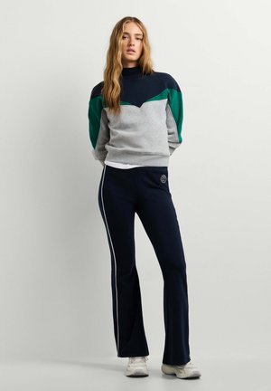 Jeune femme portant un sweat-shirt color-block et un pantalon de survêtement bleu marine avec des bandes blanches sur les côtés, debout contre un mur uni.
