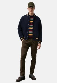 Giacca in misto lana navy con colletto a righe gialle e bianche, maglione a righe multiple, pantaloni marroni e scarpe scure. Dettagli in tessuto testurizzato.