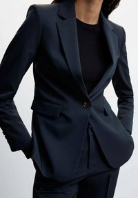 Blazer bleu marine à coupe structurée, col cranté, fermeture à un bouton, et poches latérales, associé à un haut noir côtelé ajusté.