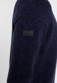 Maglione lavorato a maglia blu navy con una finitura testurizzata. Presenta un piccolo etichetta nera sulla spalla, che evidenzia la qualità del tessuto e il design.