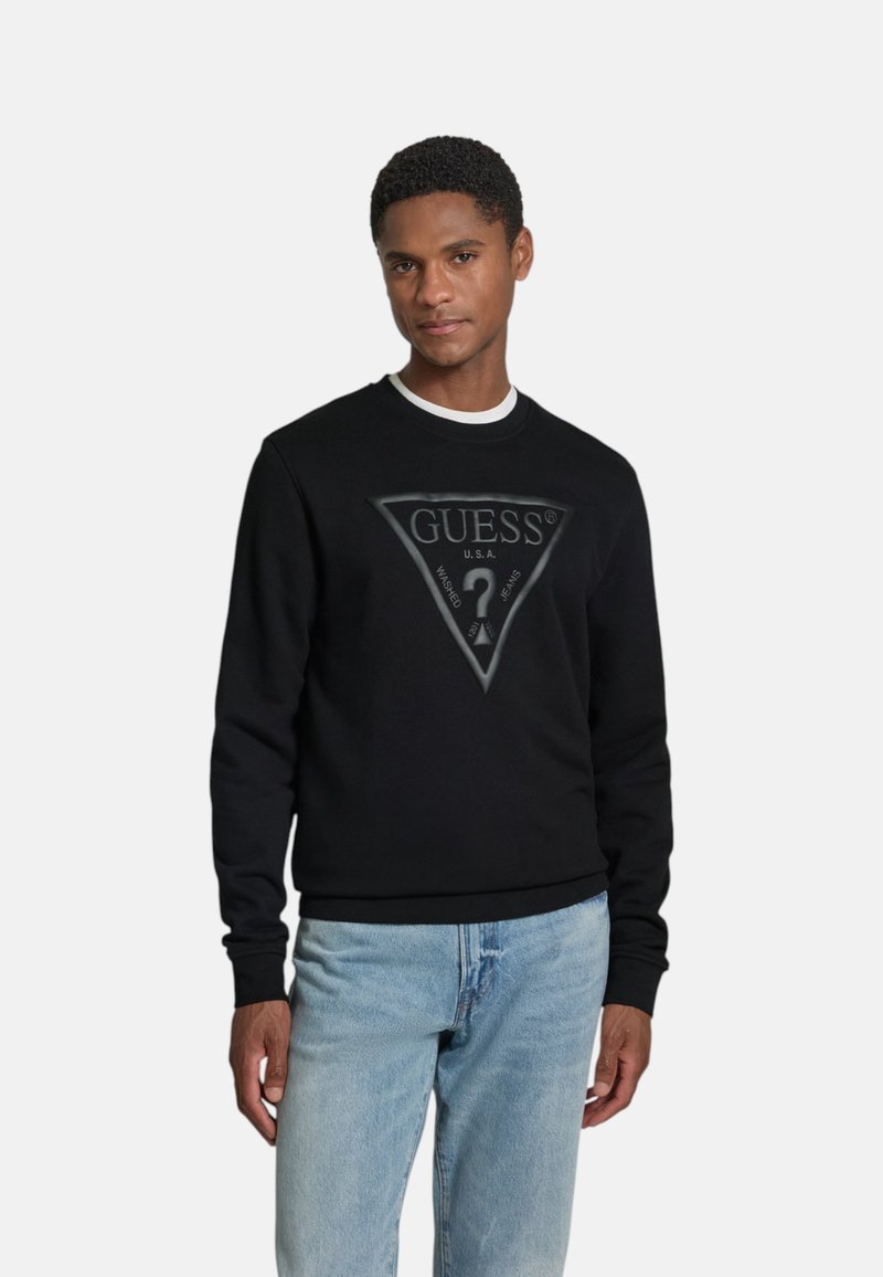 Jeune homme portant un sweat-shirt noir Guess avec un grand logo triangulaire et un jean bleu clair, debout devant un fond blanc.