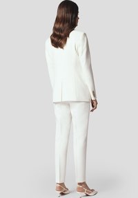 Biała dopasowana marynarka z pojedynczym wentylacją z tyłu, w zestawieniu z dopasowanymi wąskimi spodniami. Noszona z jasnymi sandałami na obcasie. Gładka tekstura materiału.