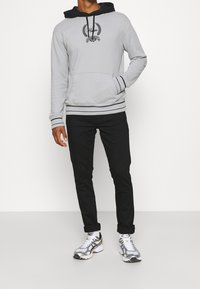 Sudadera gris con detalles en negro, bolsillo canguro y logo bordado. Combinada con pantalones negros y zapatillas deportivas plateadas.