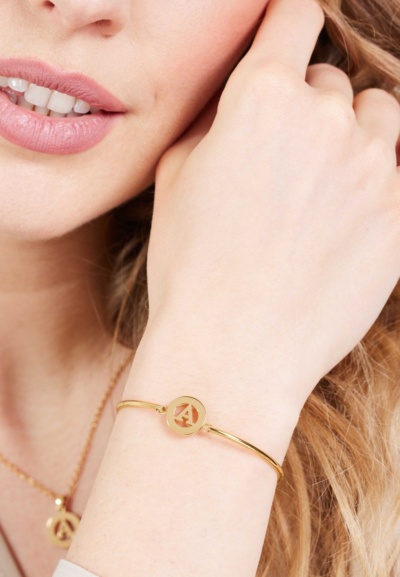 Gouden bangle armband met een ronde bedel met een uitsneden "A" logo, een gladde textuur en een strakke, minimalistische uitstraling.