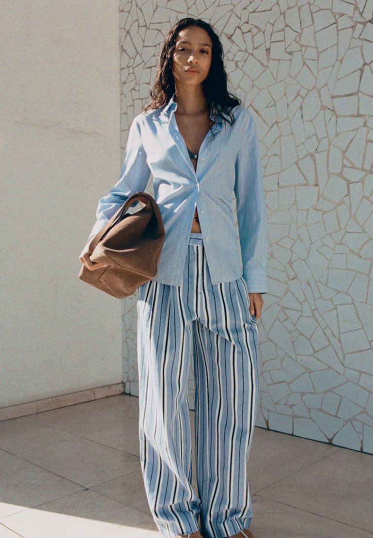 Femme en chemise bleu clair boutonnée et pantalon large rayé bleu et blanc, tenant un grand sac marron, debout devant un mur en mosaïque.