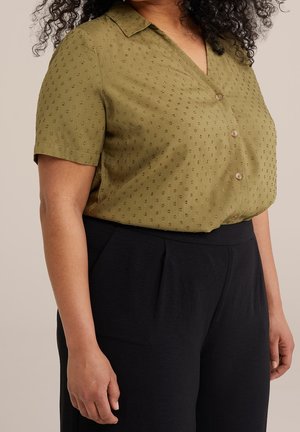 Mujer con blusa de manga corta color verde oliva con botones y puntos texturizados, y pantalones negros de talle alto, de pie frente a un fondo neutro.