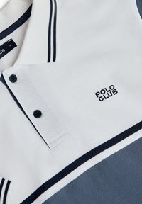 Vit polo t-shirt med marinblå ränder och krage. Har två svarta knappar och "POLO CLUB"-logotypen på bröstet i svart. Känsla av bomullstextur synlig.