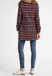 Kabát ze směsi vlny v barevném houndstooth vzoru s límcem z umělé kožešiny, detailem opasku a dlouhými rukávy, v kombinaci s džíny a stříbrnými botami.