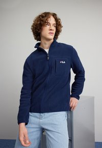 Giacca in felpa blu navy con zip frontale, colletto fitted e tasca sul petto sinistro. Presenta un logo "FILA" bianco e rosso. Superficie testurizzata.