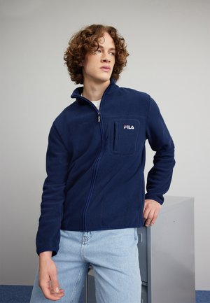Giacca in felpa blu navy con zip frontale, colletto fitted e tasca sul petto sinistro. Presenta un logo "FILA" bianco e rosso. Superficie testurizzata.