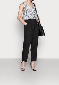 Donna che indossa un top floreale senza maniche, pantaloni neri a vita alta, sandali neri con tacco alto e tiene in mano una borsa nera.