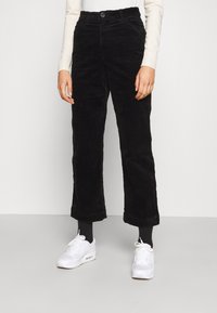 Pantalons en velours côtelé noirs à taille haute, jambe droite et ourlets retroussés, associés à des baskets blanches et un haut à manches longues léger.