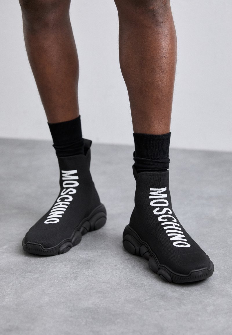 Black sock-style sneakers feature a thick, textured sole and prominent white "Moschino" logo on the sides. Made from flexible material.