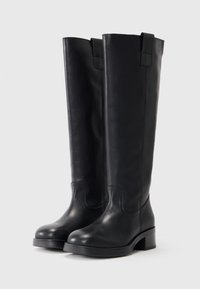Bottes en cuir noir arrivee jusqu'au genou avec une texture lisse, un bout rond et un petit talon bloc. Elles sont dotées de deux boucles en haut pour un enfilage facile.