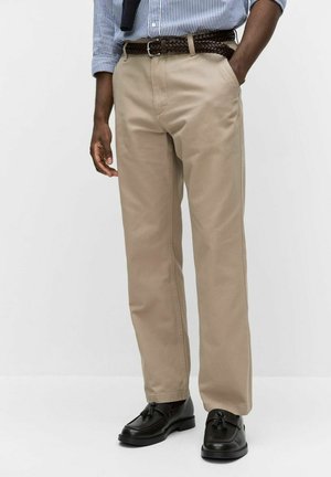 Man draagt beige broek, zwarte loafers met kwastjes, een gestreept blauw overhemd en een gevlochten bruine riem, staat met één hand in de zak.