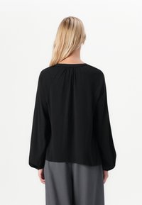 En person med blondt hår som vender bort fra kameraet, har på seg en løs, svart bluse med lange ermer og folder ved bakre yoke, samt grå, vide bukser.