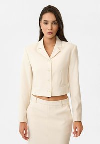 Blazer crème cintré avec un col, six boutons et des poches latérales, associé à une jupe crème assortie. Texture de tissu lisse.